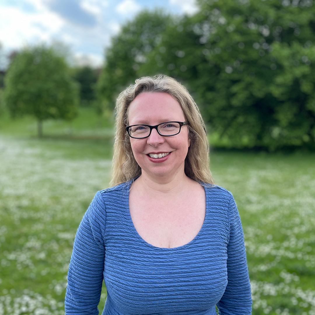 A woman with blond hair, wearing glasses and a blue top, stands in a field of daisies.