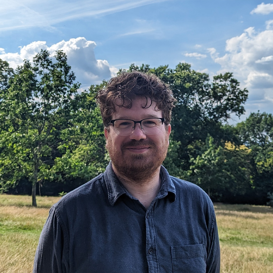 A white man, with glasses, wearing a shirt stands outside, surrounded by trees.