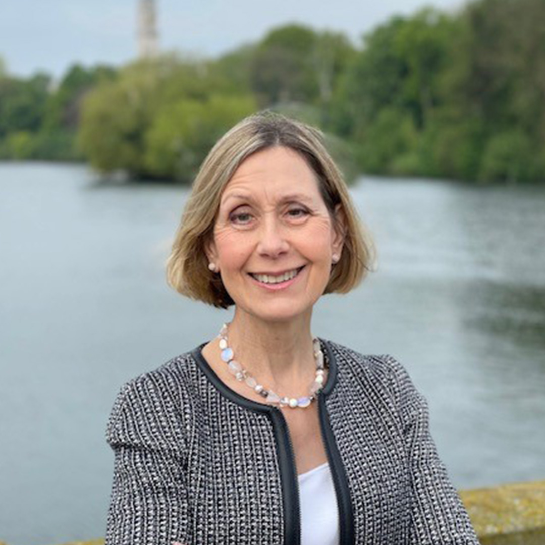A lady, with a bob haircut, wearing a smart jacket stands in front of a lake.