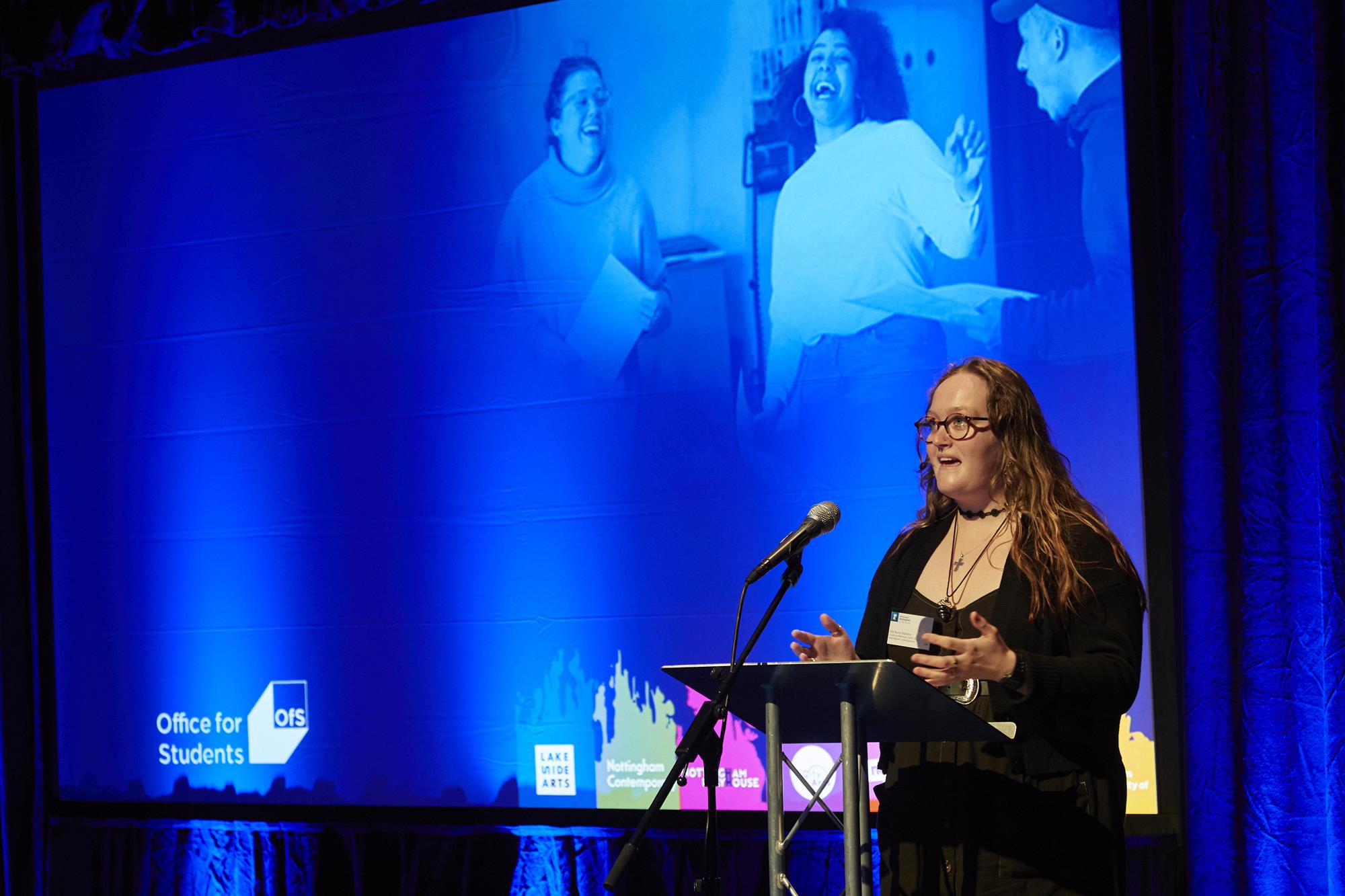 a woman presents on stage with a large blue screen behind her