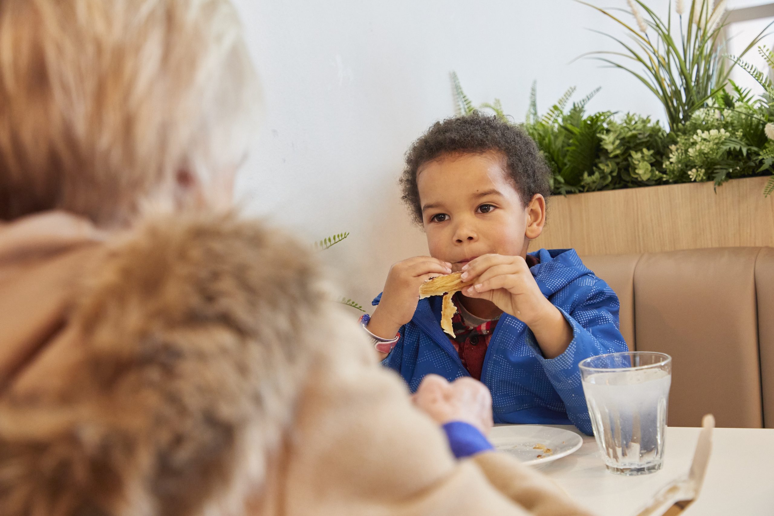 A little boy, in a blue jumper enjoys his snack at a café table.