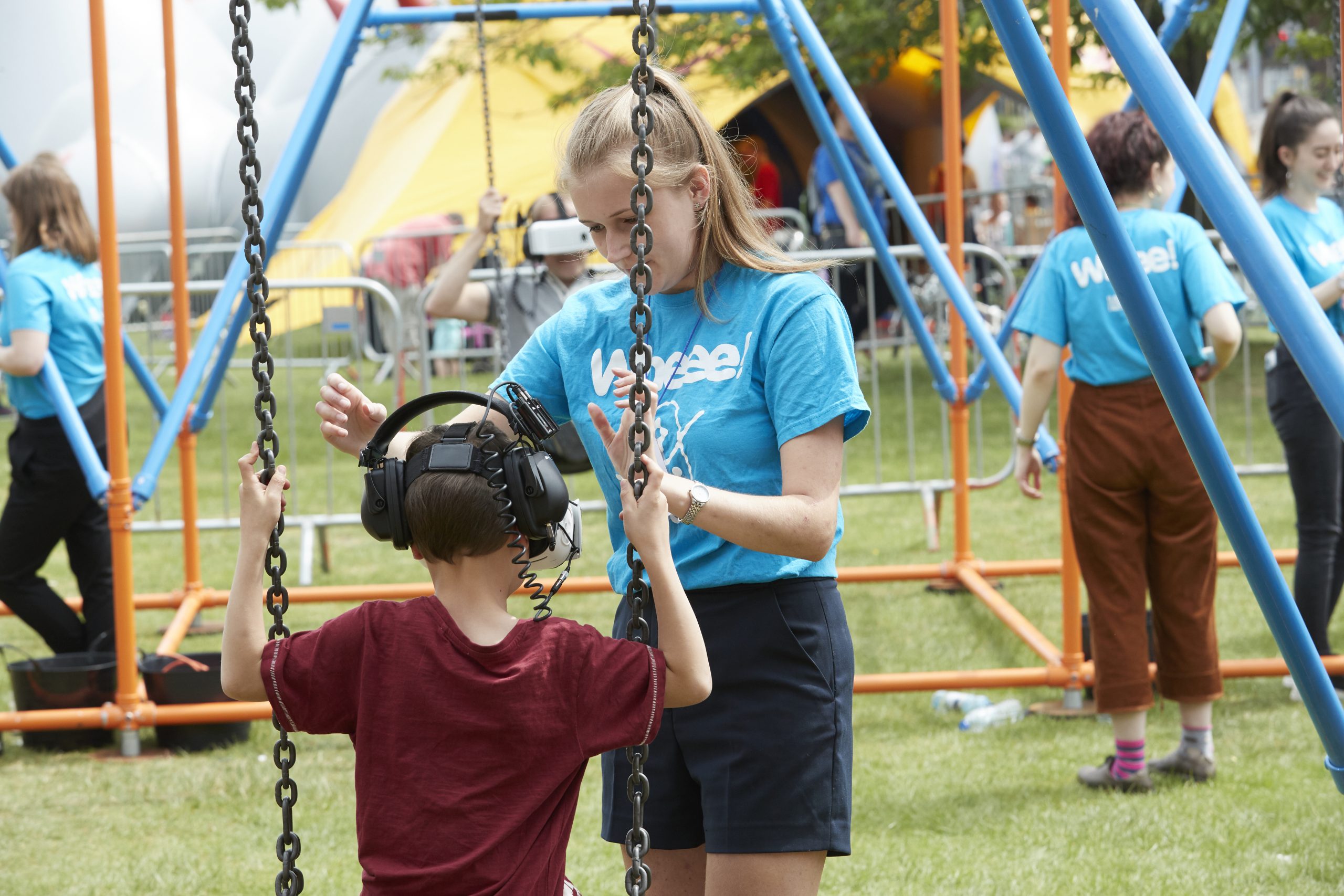 A woman in a staff t shirt helps a child on a swing in a park outside Lakeside Arts