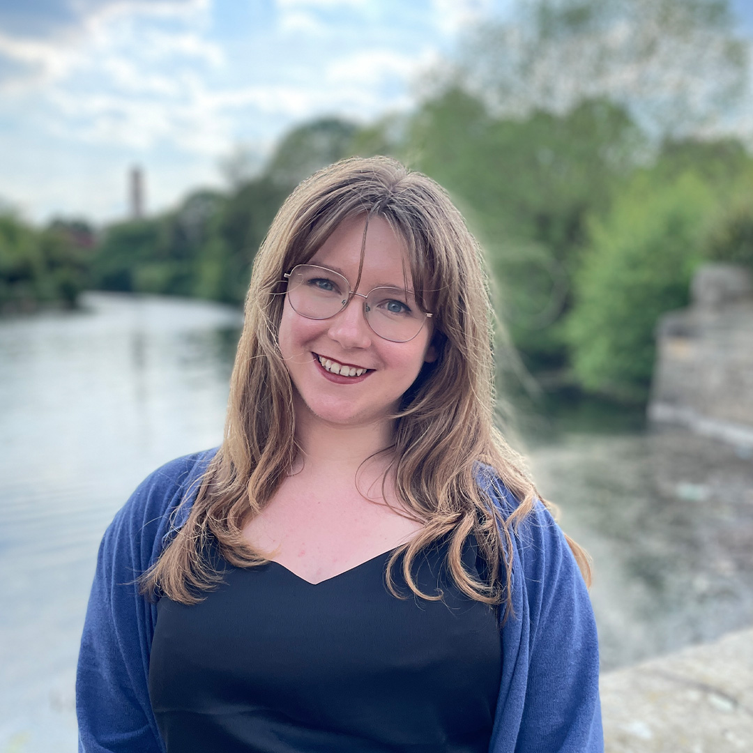 A woman with long hair and glasses stands in front of a lake wearing a blue cardigan.