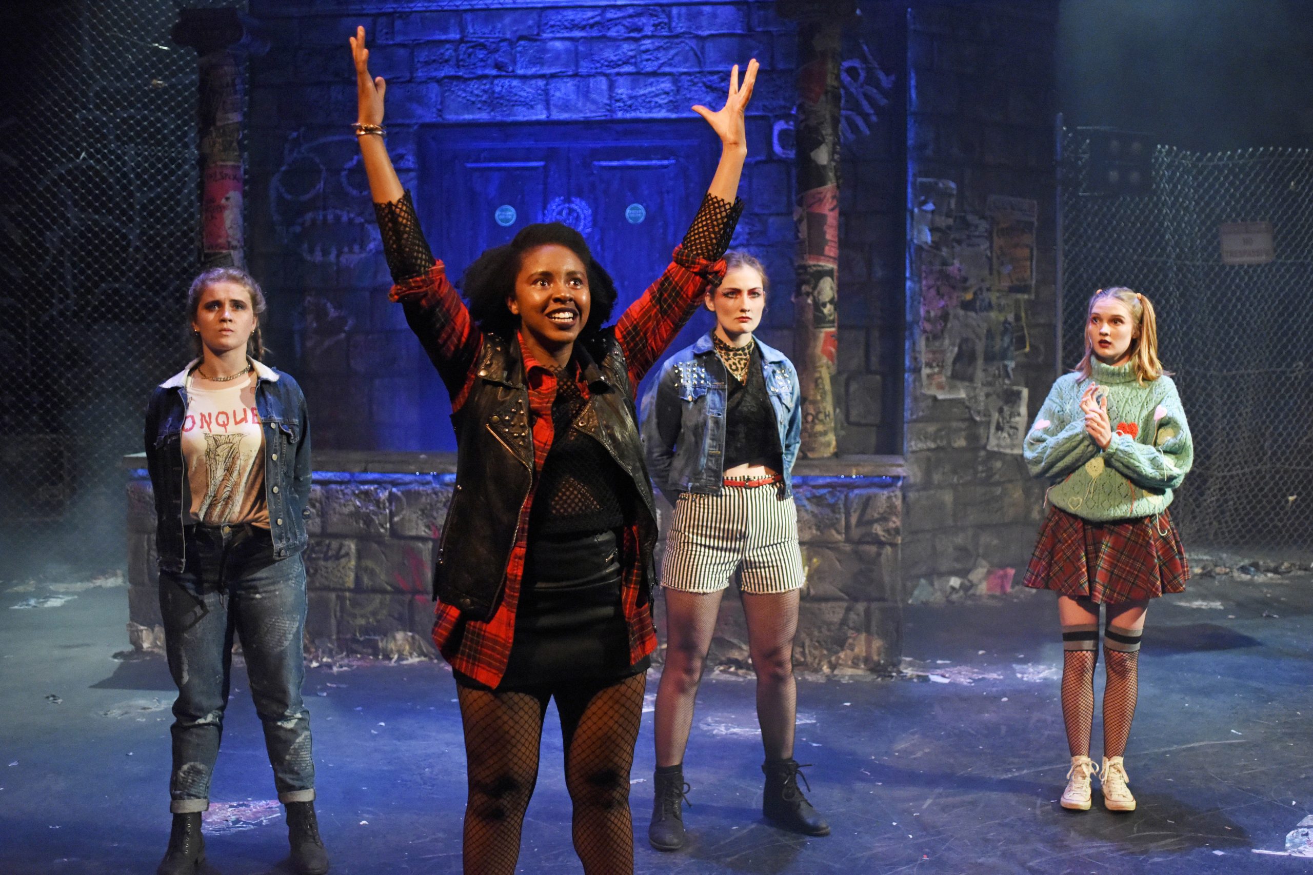 three actors stand like soldiers whilst a fourth raises her arms in glee