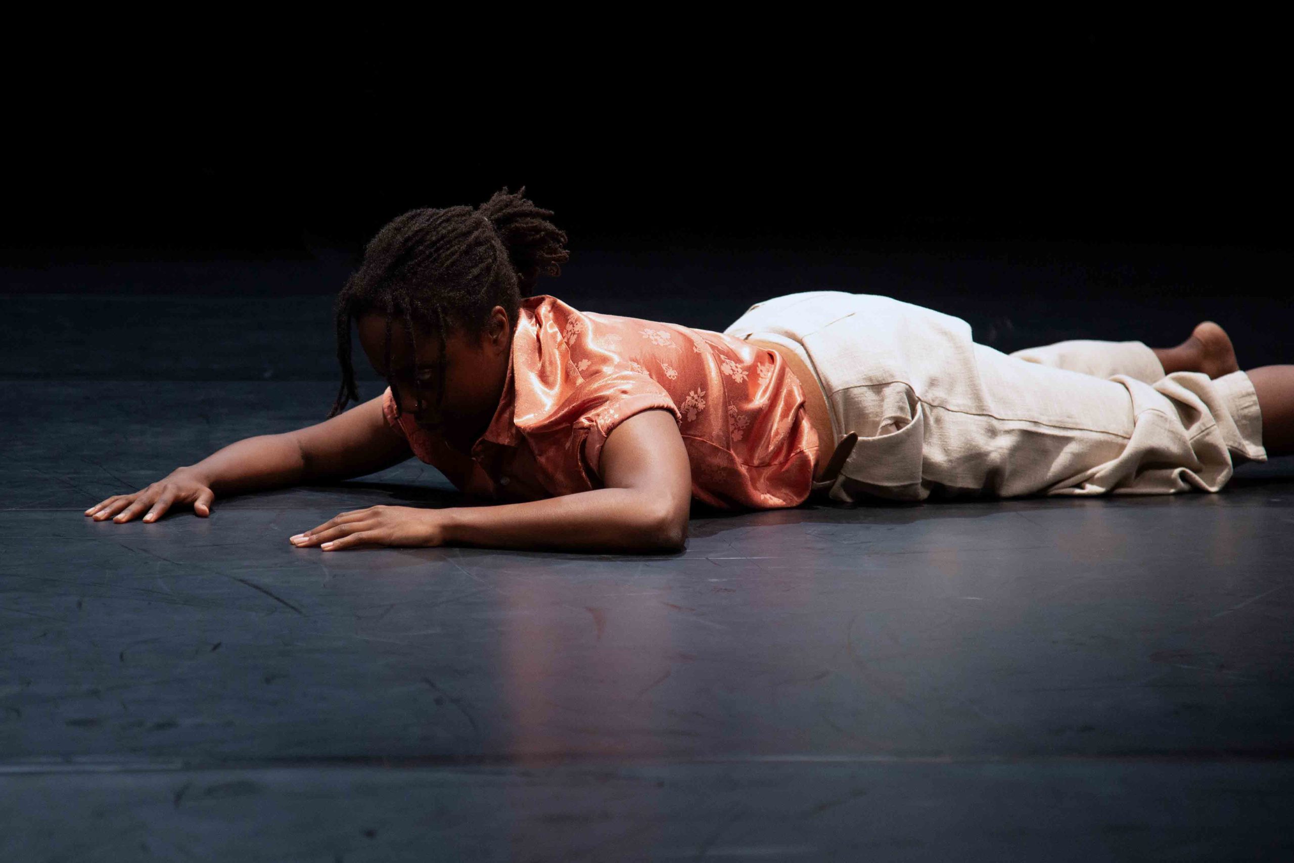 A dancer in a pink top and white trousers lies on stage