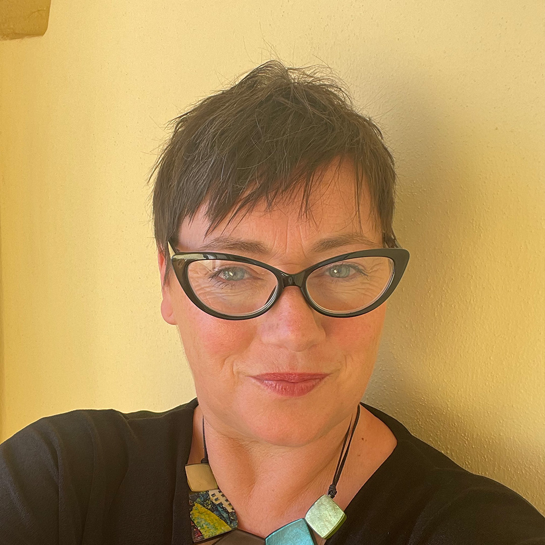 A woman with short dark hair and fabulous glasses stands against a yellow wall.