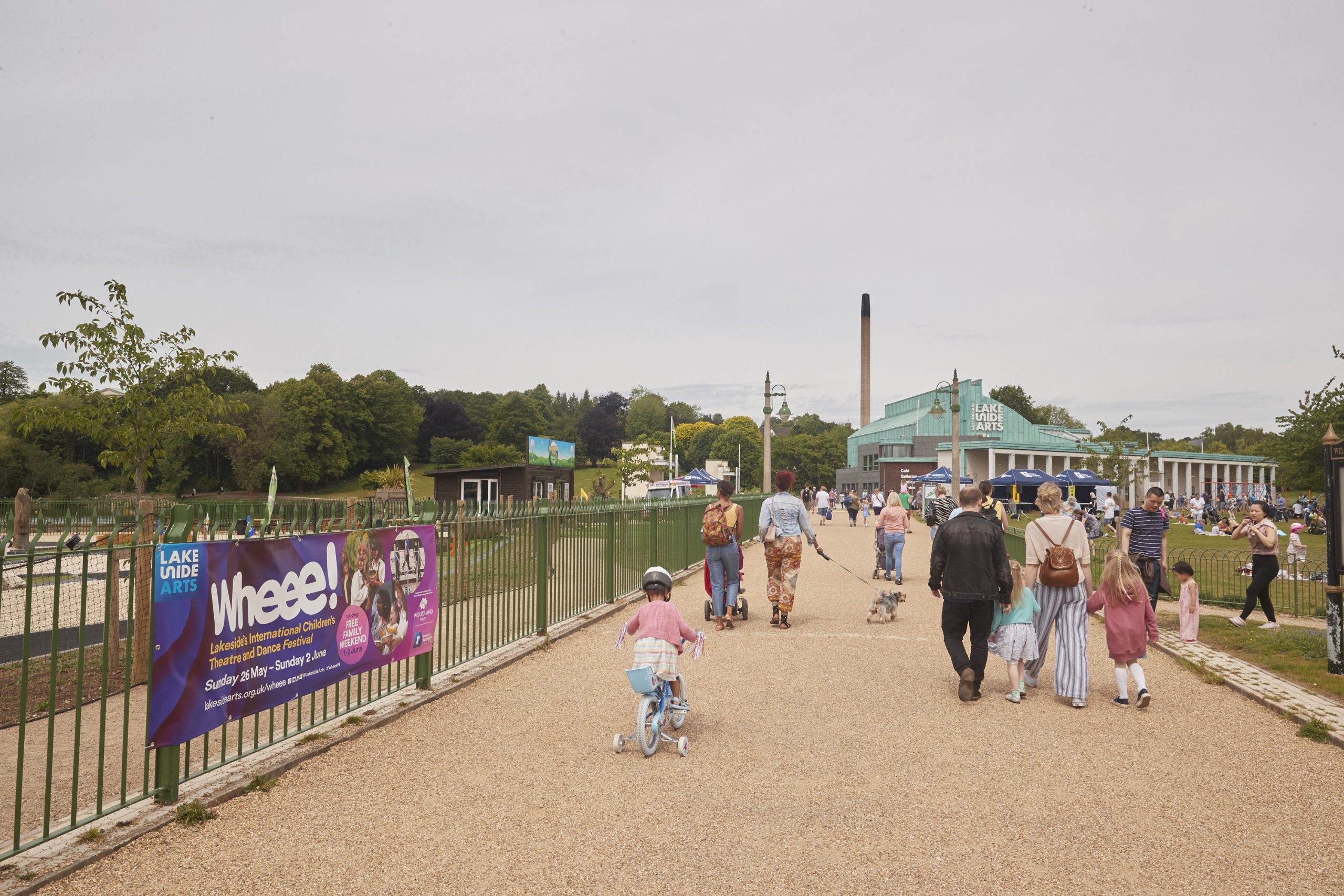 Families walk towards the Lakeside Building on a sunny day. There is a poster advertising the Wheee! Festival on a fence to the left]