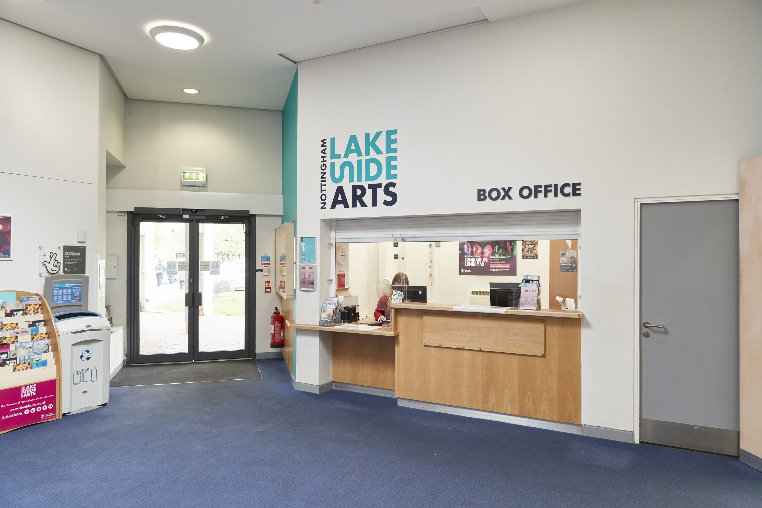 The inside of our box office, a room with a glass door, blue carpet and a desk with a woman in a red cardigan sat behind it