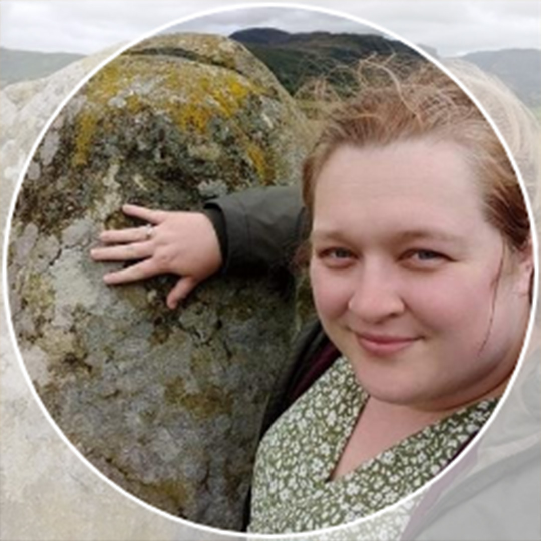 A woman with her hair tied back, in a green v neck top, stands next to a rock.
